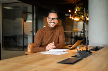 Portrait of smiling male professional in eyeglasses working over laptop at desk in modern office