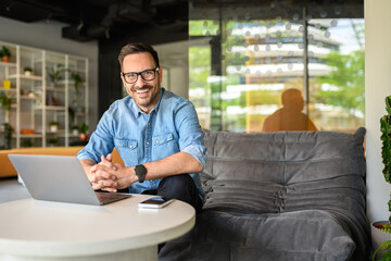 Portrait of smiling male freelancer with hands clasped sitting with laptop on sofa in modern office