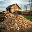 © nattapon - A wooden barn with a red roof stands in the background while a large pile of wood chips and stacked logs are in the foreground, set in a rural, pastoral landscape suitable for agriculture, forestry