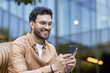 © Liubomir - Young man sitting outdoors using phone with happy expression. Smiling while engaging with smartphone, wearing glasses and casual shirt. Modern architecture in background adds urban feel to image.