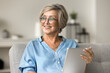 © fizkes - Smiling mature woman looking away while holding tablet seated on sofa at home, older generation and modern wireless technology and internet usage for online shopping, communication on carefree pastime