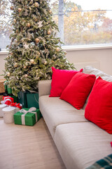 Wall Mural - Beautifully decorated Christmas tree with golden ornaments and wrapped gifts beside a beige sofa with red cushions in a bright, cozy living room.