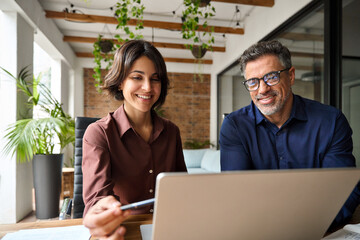 Two happy busy male and female business people working at desk using laptop computer in office. Professional business colleagues discussing corporate project idea, manager consulting client at meeting