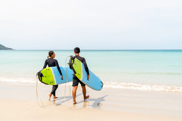 Happy Asian family couple enjoy outdoor active lifestyle water sports surfing on summer holiday vacation. Healthy people in wetsuits holding surfboard walking together on tropical beach in sunny day