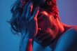 © Katavudh - A young man with a troubled expression, his hand resting on his forehead, bathed in blue and red light.