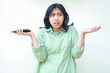 © M Alfan Setyawan - portrait of confused asian woman wearing green shirt shrug shoulders and spreads palms holding smartphone looking away worried standing over isolated background, considering gesture