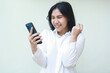 © M Alfan Setyawan - ecstatic asian business woman celebrate victory by raising fist while using smartphone wearing white shirt formal suit isolated on white background, showing yes gesture