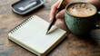 © Chonticha - A hand holding a pen, writing in a notebook with a cup of coffee beside it on a wooden desk.