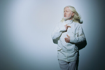 elderly man with long white hair wearing white attire against a light grey background, expressing surprise or curiosity in a minimalist setting