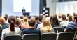 © 为轩 张 - Audience view from behind in a conference room with a white wall, watching a speaker and presentation screen. Scandinavian design style with varied colored attire and some attendees sitting back