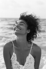 Poster - A woman with curly hair is smiling and wearing a white lace top. She is sitting on the beach and looking out at the water