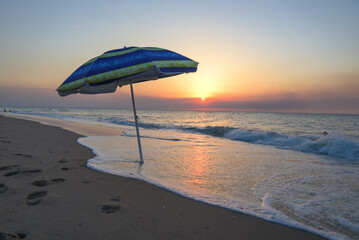 Striped beach umbrella on the beach. Beach umbrella on a sunny day, sea in background. Blue umbrella in sandy beach and in the background the mediterranean sea and sunset and waves, summer vacation.