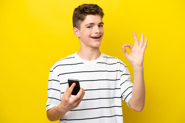 Teenager Russian man isolated on yellow background using mobile phone and doing OK sign