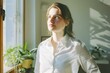 © Victor Bertrand - A woman in a white shirt stands by a sunlit window, basking in natural light with a thoughtful expression, surrounded by indoor plants.