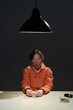 © pressmaster - Man in orange jumpsuit with glasses sitting under hanging lamp with focused expression, holding small item on table with dark backdrop. Setting suggests an interrogation scene