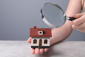 Wall Mural - House hunting. Woman with magnifying glass and house model at grey table, closeup