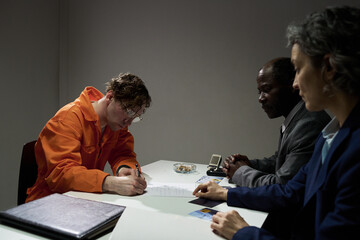 In a dimly lit room, one person in orange uniform is writing, observed by two individuals from different backgrounds wearing suits sitting close by