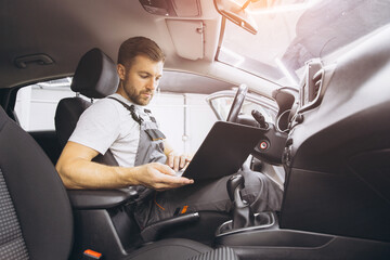 Wall Mural - Automotive mechanic running diagnostics software on computer. Mechanic sitting in car and working on laptop while doing vehicle diagnostic test in auto service garage.