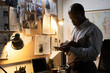 © pressmaster - Middle-aged African American man attentively examining notes on bulletin board under soft lamp light, surrounded by office supplies in a room with subdued lighting
