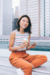 © BullRun - Portrait of cheerful hipster girl with cellular gadget in hand smiling at camera while resting at urban setting in metropolis, positive Asian female blogger with mobile phone and coffee to go