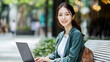 © pittari - A young and beautiful Korean woman in her late twenties is sitting on a bench outside with a laptop, smiling at the camera. She has long hair and dress in casual attire.
