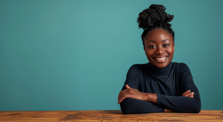 Wall Mural - African woman with a black shirt and dreadlocks is smiling at the camera. She is sitting at a wooden table with her arms crossed