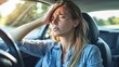 © Ganesha - A tired woman is sitting in the driver's seat of her car, with one hand on her forehead, and has a stressed expression with her eyes closed. Her face shows the stress of driving