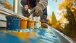 © Lamina - Homeowner painting gutters to match the house trim, with paint cans and brushes scattered on the ground below, enhancing the home’s exterior appearance and cohesion.