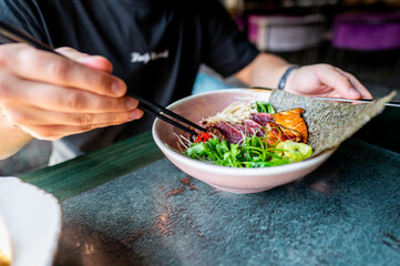 A person enjoys a bowl of Asian noodle soup with chopsticks, featuring vibrant ingredients like green onions and meat. The scene captures the essence of traditional Asian cuisine in a cozy setting.