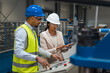 © 24K-Production - Female Indian factory supervisor is on-the-job training an African American male employee, in a blue helmet and yellow vest, to operate machinery.