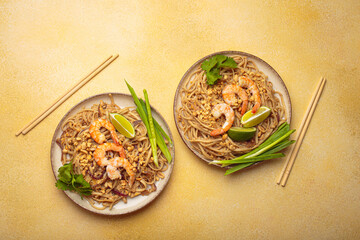 Two plates of pad thai noodles garnished with shrimps, lime, peanuts and chives, accompanied by chopsticks on a yellow background. Thai cuisine