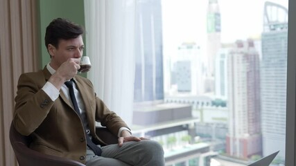 Wall Mural - Businessman sitting on furniture drinking coffee while working on laptop at ornamented corporate waiting area for meeting preparation with overlooking window of cityscape skyline background