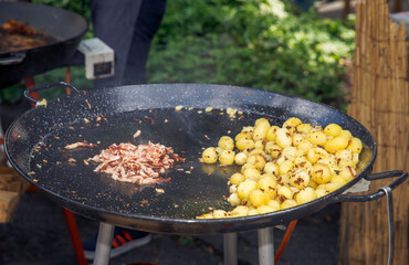 Outdoor cooking, fresh roasted potatoes and bacon on large black fry pan, nature background, food preparation concept