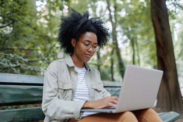 Black woman focused on laptop work, enjoying a summer day on a park bench