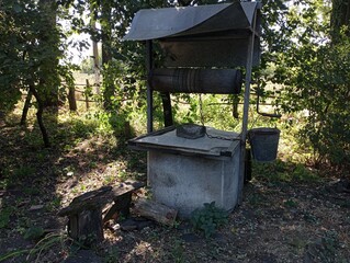 An old picturesque well with water in the middle of the yard in the shade of tall trees made of concrete rings with a metal roof and a bucket attached to the side. Water source in the village. Old vil