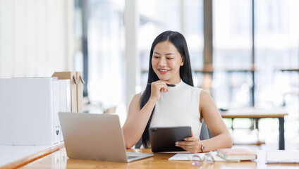 Wall Mural - Positive young asian female freelancer in formal suit sitting at table with laptop and browsing tablet while working on project in creative workspace in daylight