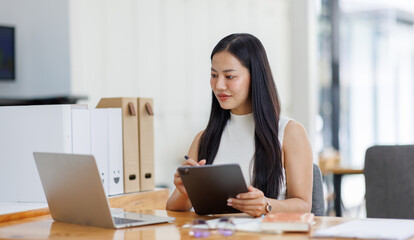 Wall Mural - Positive young asian female freelancer in formal suit sitting at table with laptop and browsing tablet while working on project in creative workspace in daylight