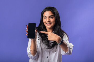 A young woman of Indian descent, wearing a white striped shirt and black smartwatch, stands against a solid purple background, smiling and extending a smartphone forward, presenting it