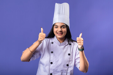 A cheerful Indian woman in a purple chef coat and white chef hat gives two thumbs up, standing against a solid purple background, portraying confidence and positivity in her culinary profession