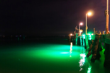 A serene nighttime view of a glowing pier illuminated by green lights, reflecting on calm water under a dark sky.