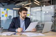 © Liubomir - Businessman in formal suit focused on paperwork at office desk. Concentrated on financial documents, using pen and laptop for detailed analysis. Modern workspace reflects professional environment.