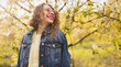 © kegfire - A joyful woman wearing a denim jacket and sunglasses smiles while enjoying a sunny spring day outdoors in nature with blooming trees.