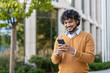 © Liubomir - Young professional with curly hair using smartphone outdoors. Scene conveys connectivity, technology, and communication. Individual dressed in casual autumn attire, standing confidently near office