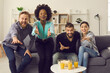 © Studio Romantic - Multiethnic people watching sports game sitting on sofa at home. Group of multiracial friends enjoying soccer match on TV, supporting players and waiting for goal that brings victory to favorite team