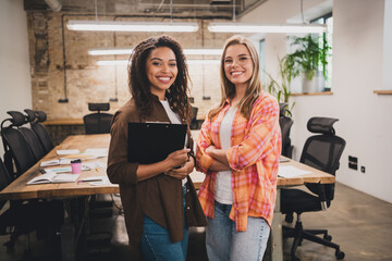 Portrait of two young corporate girls clipboard crossed hands coworking loft interior business center office indoors