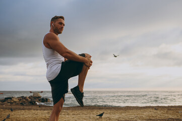 Wall Mural - Side view young strong sporty toned fit sportsman man wear sports clothes earphones raise up leg hold knee warm up training at sunrise sun dawn over sea beach outdoor on seaside in summer day morning.