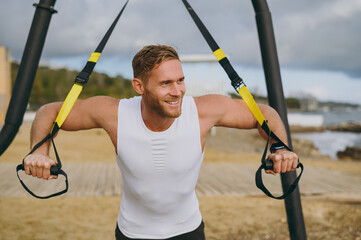 Wall Mural - Young sporty athletic toned fit sportsman man he wearing sports clothes use trx loops do exercise look aside warm up train at sunrise sun dawn over sea beach outdoor on seaside in summer day morning.