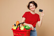 © ViDi Studio - Young woman wear red t-shirt casual clothes hold basket bag with food products, mock up of credit bank card isolated on plain pastel light beige background. Delivery service from shop or restaurant.