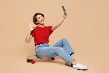 Wall Mural - Full body young woman wear red t-shirt casual clothes sit on pennyboard skateboard doing selfie shot on mobile cell phone post photo on social network show v-sign isolated on plain beige background.