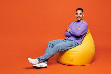 Full body young happy man of African American ethnicity he wear purple sweatshirt casual clothes sit in bag chair hold hands crossed folded isolated on plain red orange background. Lifestyle concept.